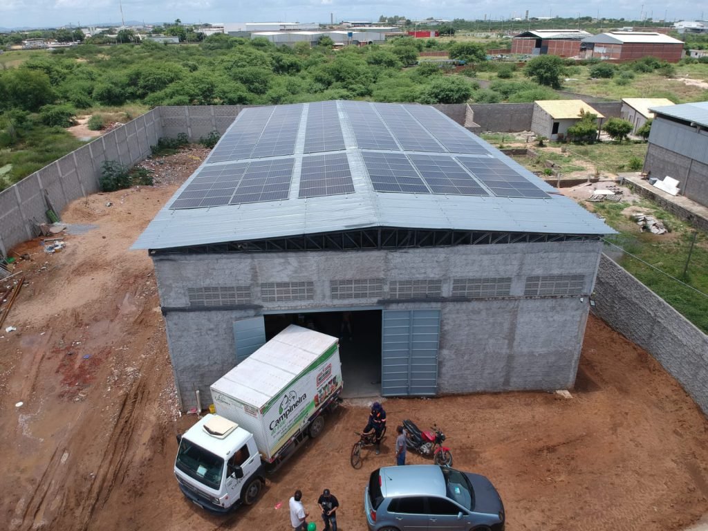 Vista aérea de um sistema fotovoltaico comercial da Senso Solar em Petrolina, com painéis solares cobrindo o telhado de um galpão industrial e céu claro ao fundo.